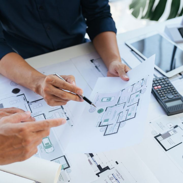 A close-up shot of two people's hands reviewing architectural blueprints on a white desk. One person points to a detail on a floor plan while the other holds a pen over another section. A tablet, calculator, and coffee cup are also on the desk.