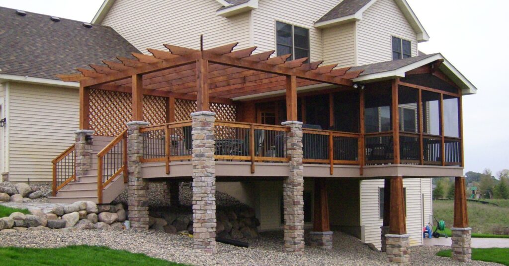 A large, elevated deck featuring a substantial wooden pergola with stone columns, attached to a beige-sided house with a gravel and rock landscape below.