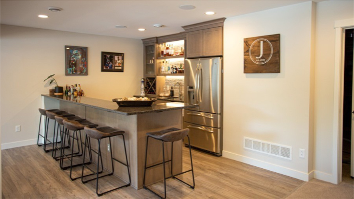 A stylish finished basement featuring a wet bar with a concrete-style countertop, wood-laminate flooring, and modern black metal bar stools. The area includes a stainless steel refrigerator, backlit shelving for glassware, and contemporary wall art.