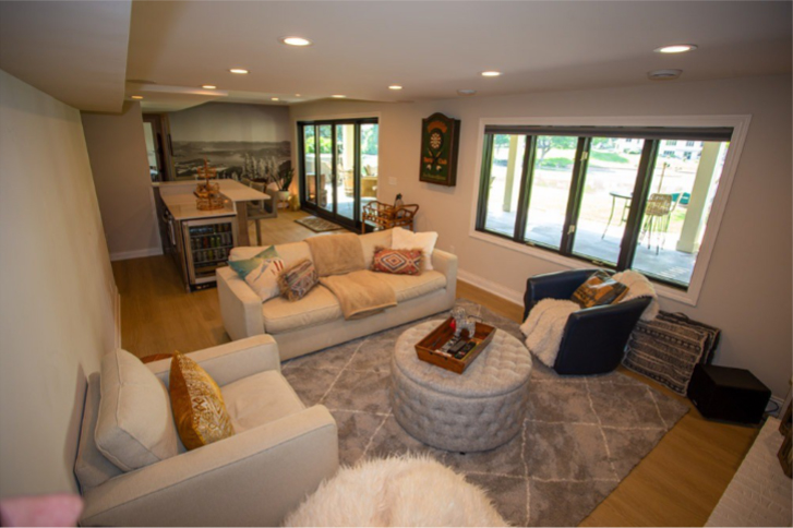 A well-lit, finished basement living space featuring light-colored furniture, including sofas and armchairs, arranged on a large area rug. In the background, there is a small kitchenette and large windows looking out to a patio.