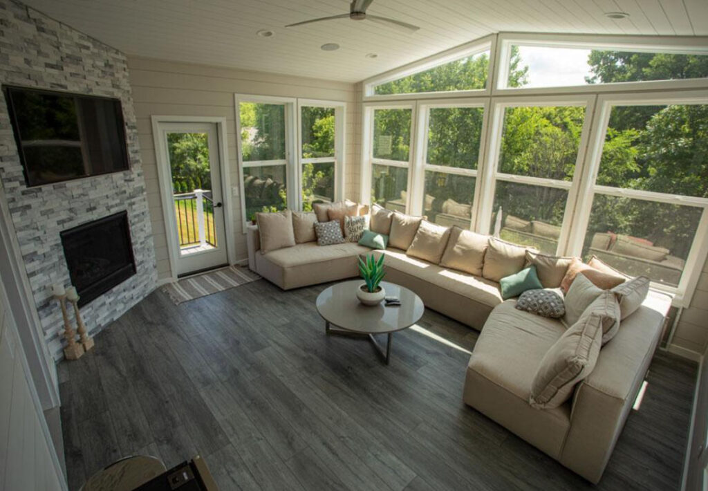 A bright sunroom with an L-shaped beige sofa, a stone fireplace with a mounted TV, and large windows offering a view of lush green trees outside.