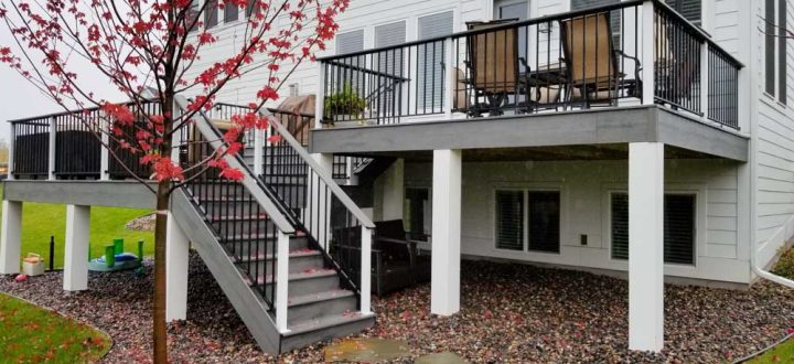 A two-tiered gray deck with black railings attached to a white-sided house, featuring a red maple tree in the foreground and a gravel landscape below.