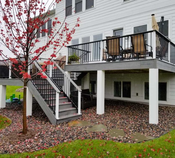 A two-tiered gray deck with black railings attached to a white-sided house, featuring a red maple tree in the foreground and a gravel landscape below.
