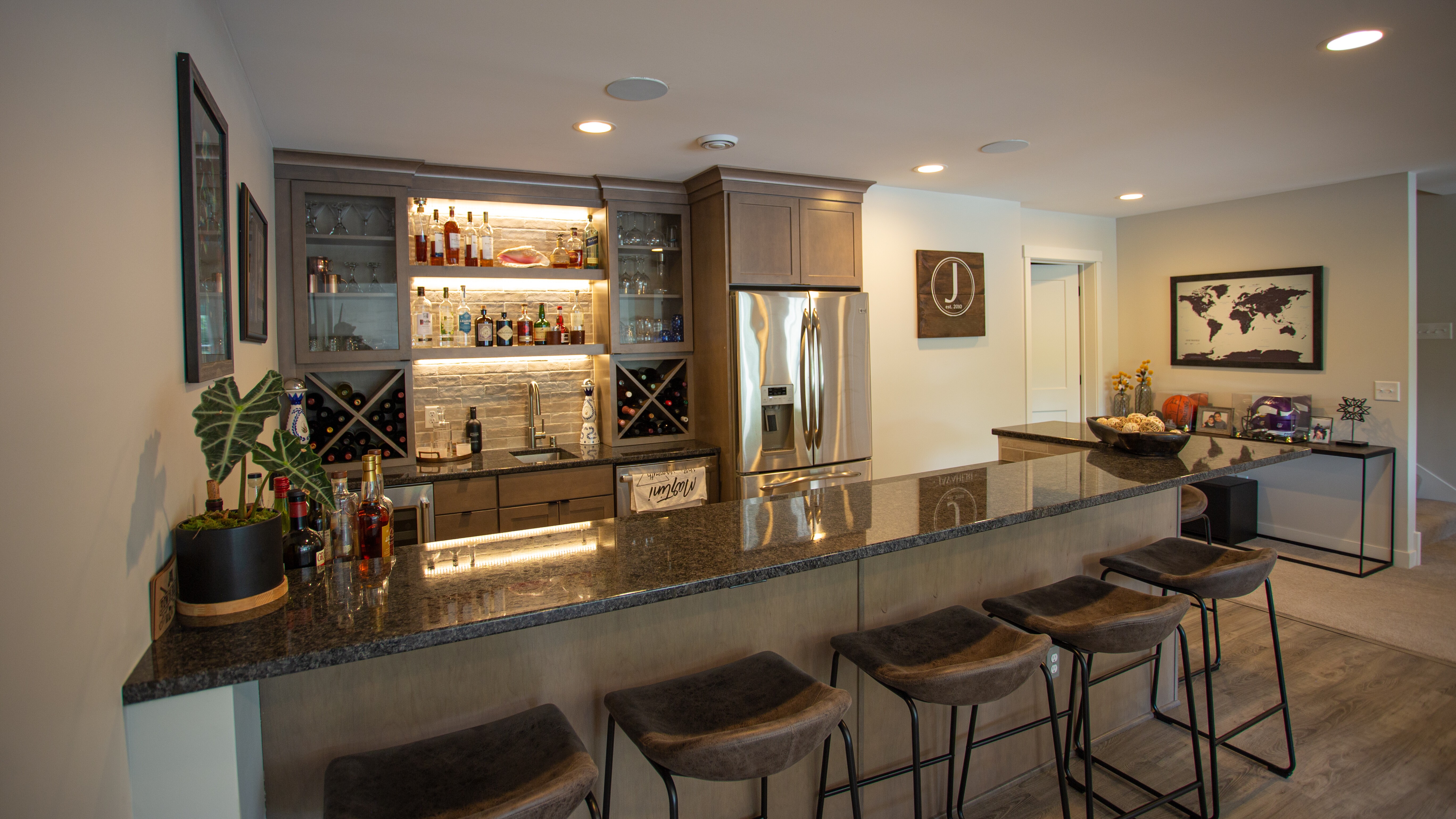 A modern home bar with backlit shelves displaying bottles, a stainless steel refrigerator, a granite countertop, and bar stools.