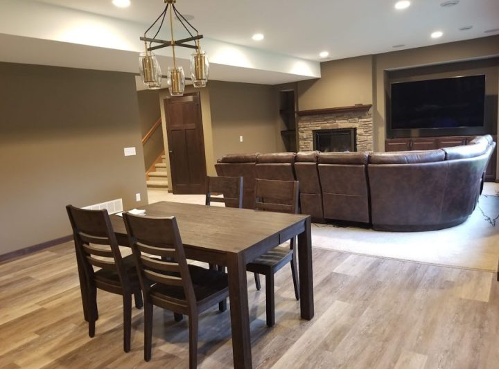 A finished basement featuring a dark wood dining table, a stone fireplace, a large curved sectional sofa for a home theater, and recessed lighting.
