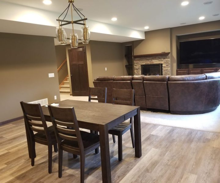 A finished basement featuring a dark wood dining table, a stone fireplace, a large curved sectional sofa for a home theater, and recessed lighting.