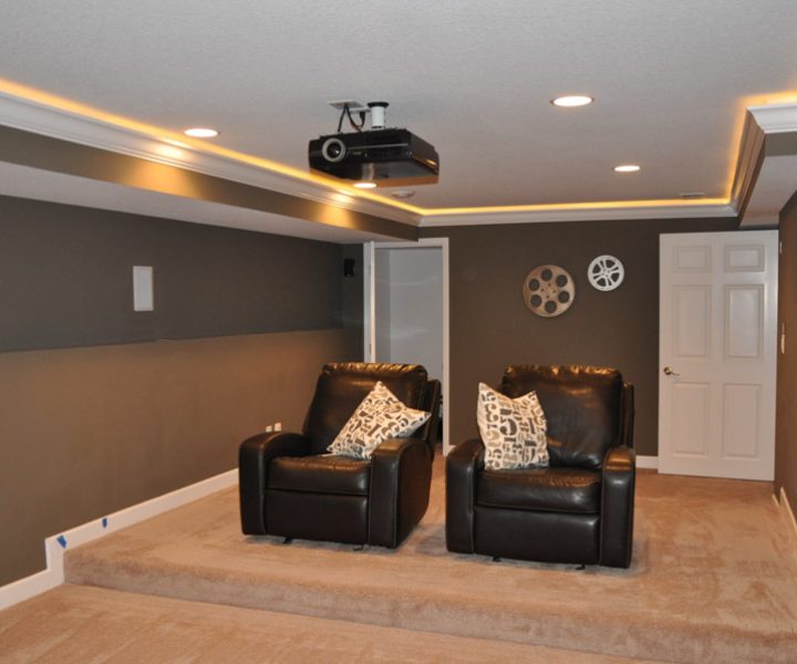 A dedicated home theater room with dark brown walls, two black leather recliners, a ceiling-mounted projector, and ambient lighting.
