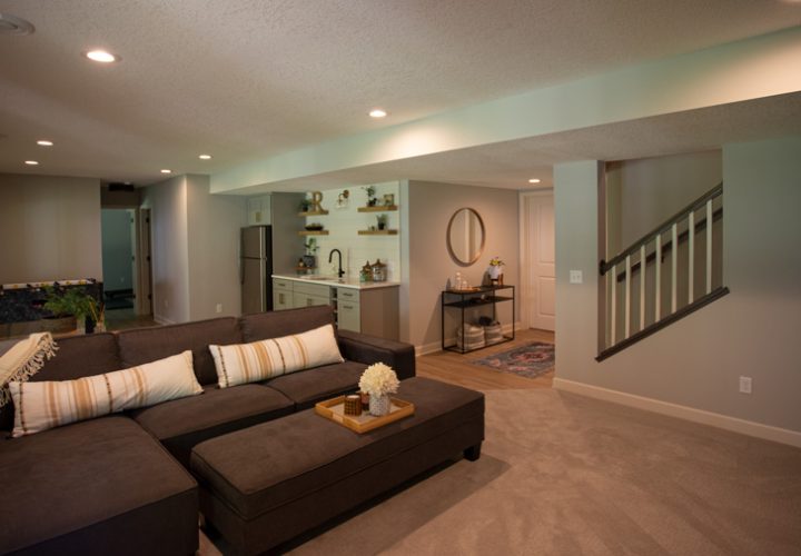 A cozy basement living room featuring a large brown sectional sofa, a wet bar with open shelving, and a view of the staircase leading to the upper level.