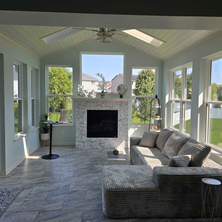 A spacious sunroom featuring a stone fireplace, light blue walls, vaulted ceiling with skylights, and large windows offering views of a green backyard.