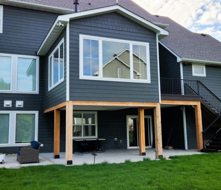 The back of a modern dark gray house with large windows, an open lower patio area, and a black metal staircase leading up to the deck.