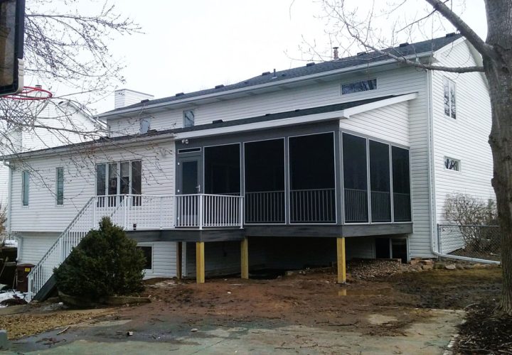 A white-sided home featuring a newly built gray-screened porch with white railings, set against a backdrop of bare trees and a muddy yard.