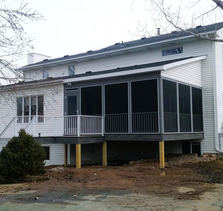 A white-sided home featuring a newly built gray-screened porch with white railings, set against a backdrop of bare trees and a muddy yard.