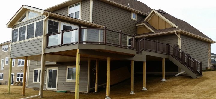 A substantial two-tiered deck with brown railings and stairs attached to a gray-sided house, situated on a sloped, dry grassy yard.