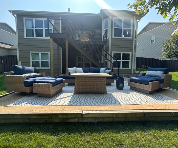 A lower-level patio with wicker furniture and a fire pit table on a patterned rug, situated in front of a gray home with a modern upper deck and stairs.