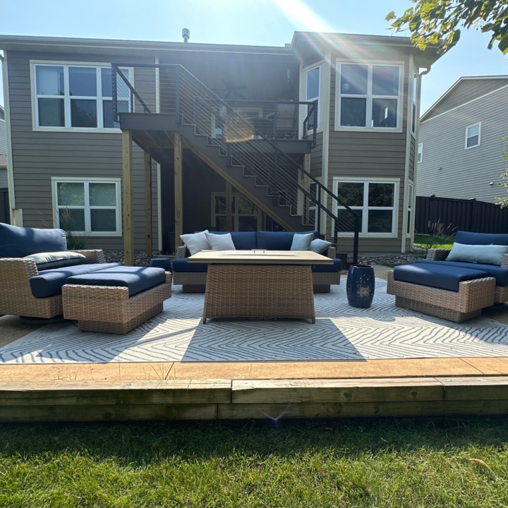A lower-level patio with wicker furniture and a fire pit table on a patterned rug, situated in front of a gray home with a modern upper deck and stairs.