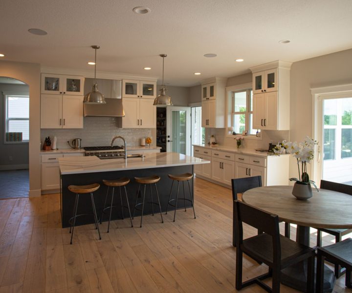 A bright, modern kitchen featuring white cabinets, a dark island with bar stools, stainless steel appliances, and hardwood floors, opening to a dining area.