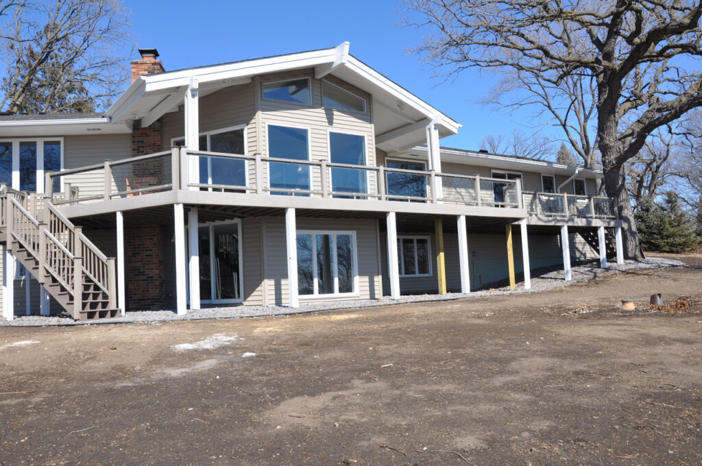 The back of a beige home with a large, elevated deck featuring glass railings, supported by white posts, set against a backdrop of bare trees and a dirt yard.