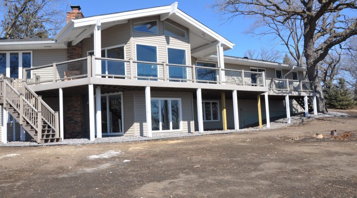 The back of a beige home with a large, elevated deck featuring glass railings, supported by white posts, set against a backdrop of bare trees and a dirt yard.