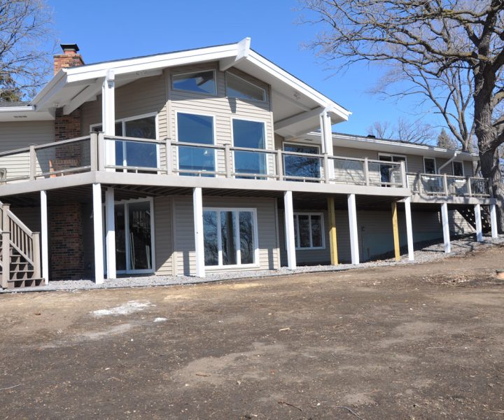 The back of a beige home with a large, elevated deck featuring glass railings, supported by white posts, set against a backdrop of bare trees and a dirt yard.