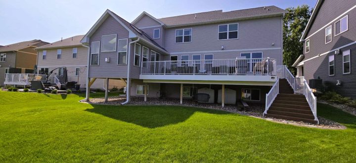 A large, light gray two-story home with a white-railed upper deck and stairs, set against a vibrant green backyard under a clear blue sky.