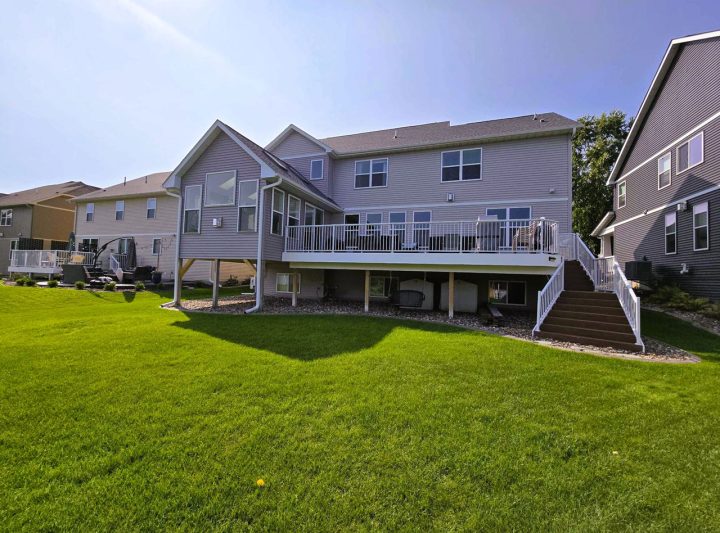 A large, light gray two-story home with a white-railed upper deck and stairs, set against a vibrant green backyard under a clear blue sky.