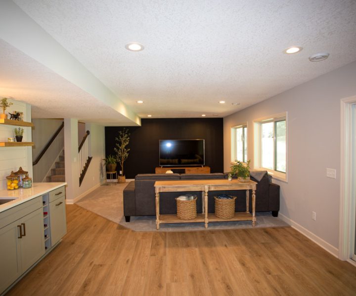 A finished basement featuring a wet bar with white cabinets, a large TV on a black accent wall, a gray sectional sofa, and a sliding glass door to the backyard.