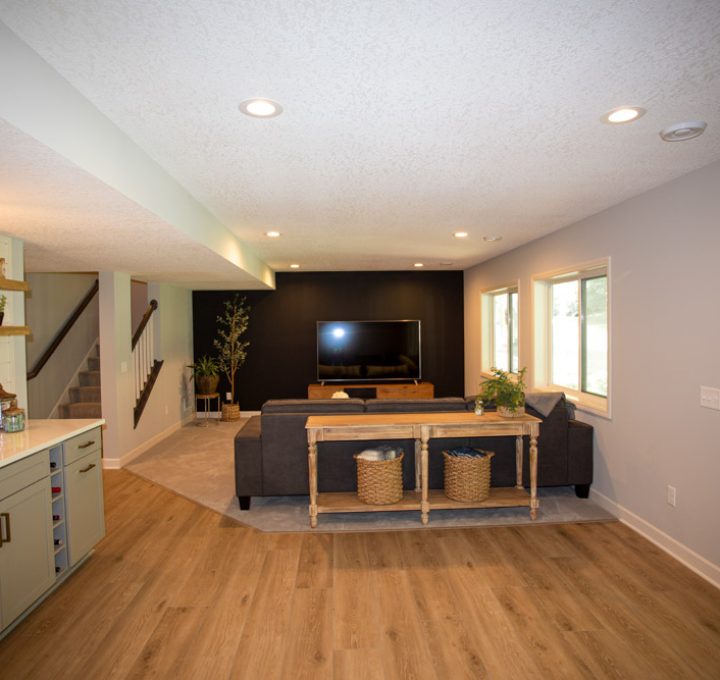 A finished basement featuring a wet bar with white cabinets, a large TV on a black accent wall, a gray sectional sofa, and a sliding glass door to the backyard.