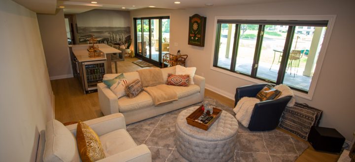 A cozy basement living room featuring a light-colored sectional sofa, round tufted ottomans, a dartboard on the wall, and large windows overlooking a patio.
