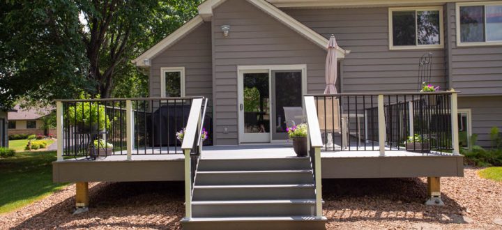 A gray-sided home featuring a modern, elevated deck with black railings and stairs, set against a landscaped yard with mulch and green grass.