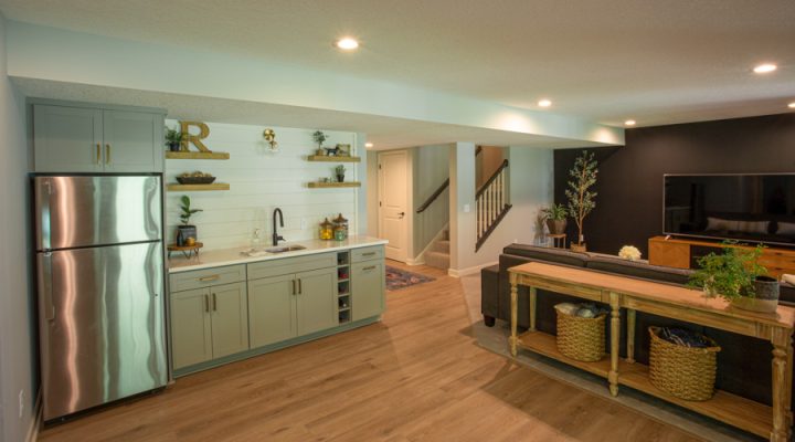 A modern, open-plan kitchen featuring a refrigerator, grey cabinetry, and wooden accents with a nearby entertainment area with a flat-screen TV and dark accent wall.