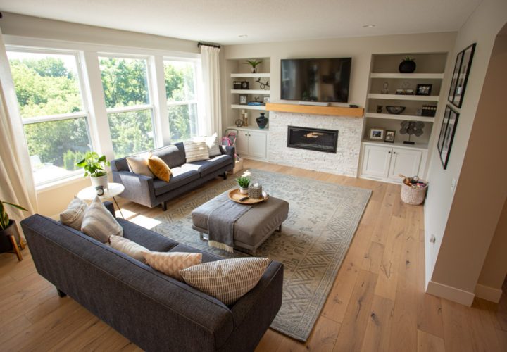 A beautifully designed family room with a stone fireplace, built-in shelving, and plush seating, highlighted by large windows and warm wooden flooring.