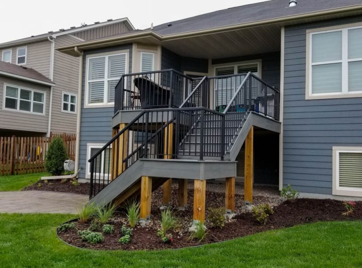 A two-tiered gray deck with black railings attached to a blue-sided house, situated on a landscaped yard with mulch and small plants.