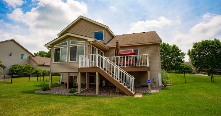 A view of a suburban home's back exterior showing an elevated deck with stairs leading down to a well-maintained lawn, surrounded by a privacy fence.