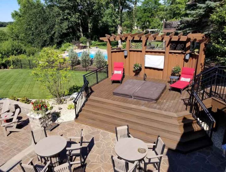 A multi-level wooden deck featuring a pergola, a lounge area with red chaise lounges, and a view of a swimming pool and green lawn beyond a black fence.