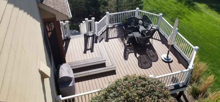 An elevated deck with white railings and stairs, featuring outdoor furniture and an umbrella, overlooking a steep, grassy slope and trees.