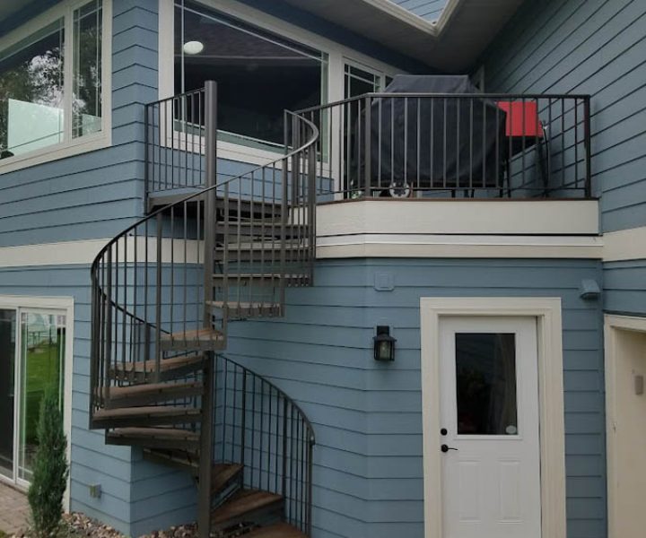 A blue-sided home featuring a spiral staircase with black railings leading up to an upper-level deck with outdoor furniture.