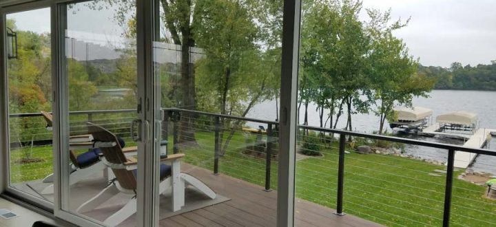 A view from inside looking out through large sliding glass doors to a wooden deck with cable railings, overlooking a green lawn and a lake with docks.