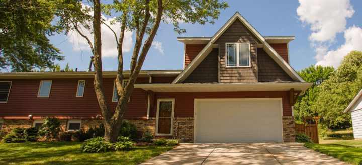 A wide-angle shot of a two-story home's exterior on a sunny day. The house features red-brown siding, stone accents, and a prominent gable over a white garage door. A concrete driveway leads up to the garage, framed by a green lawn and mature trees.