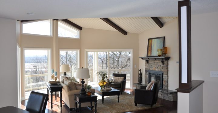 A spacious living room featuring a stone fireplace, a vaulted ceiling with exposed beams, hardwood floors, and large windows offering a scenic view.