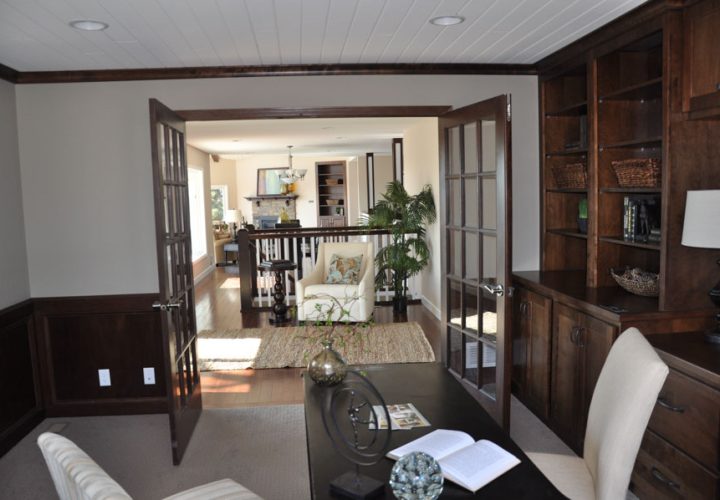 A home office featuring built-in dark wood bookshelves, a black desk, and French doors opening to a living area with a view of the fireplace.