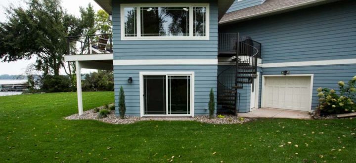 The side view of a blue-sided home featuring a spiral staircase leading to an upper deck, a lower-level sliding glass door, and a view of a lake beyond the green lawn.