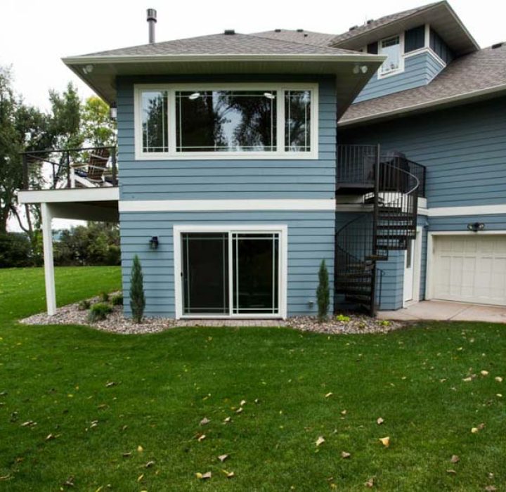 The side view of a blue-sided home featuring a spiral staircase leading to an upper deck, a lower-level sliding glass door, and a view of a lake beyond the green lawn.