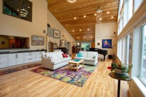 An open-concept living room with a high vaulted wood ceiling, hardwood floors, white sofas, a colorful area rug, and a view into the kitchen.
