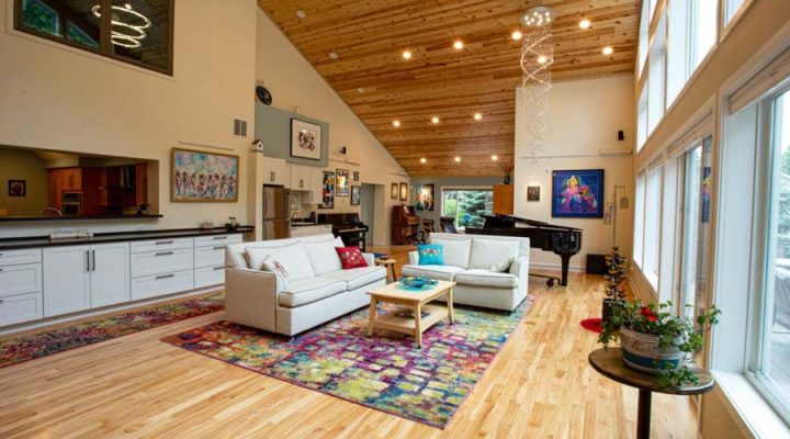An open-concept living room with a high vaulted wood ceiling, hardwood floors, white sofas, a colorful area rug, and a view into the kitchen.