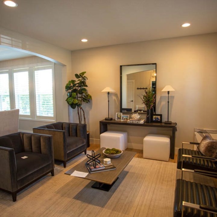 A modern foyer featuring two gray armchairs, a dark console table with a mirror, white ottomans, and large windows with white shutters.