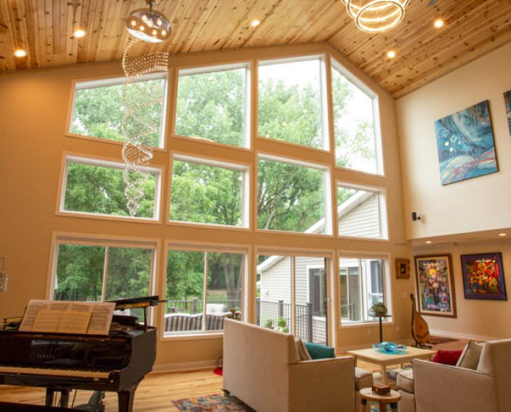 A sunlit living room featuring a high vaulted wood ceiling, large windows offering a view of trees, a grand piano, and modern art on the walls.