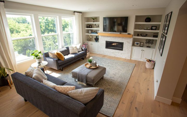A bright living room with hardwood floors, a white brick fireplace, built-in shelves, and large windows offering a view of green trees outside.