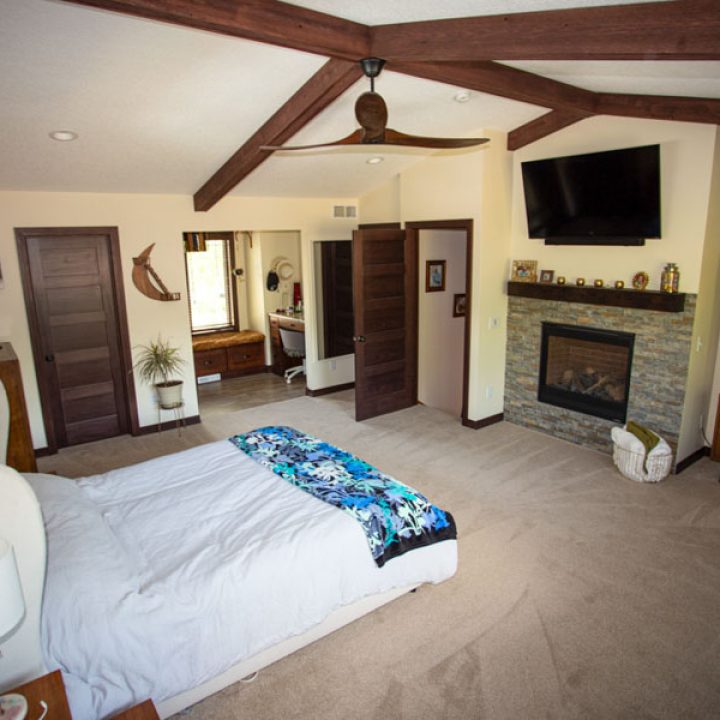 A spacious master bedroom featuring a vaulted ceiling with exposed beams, a stone fireplace, a mounted TV, and a large bed with white bedding.