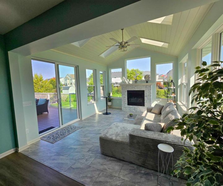 A bright sunroom with light blue walls, a stone fireplace, vaulted ceiling with skylights, and large sliding glass doors opening to a wooden deck.