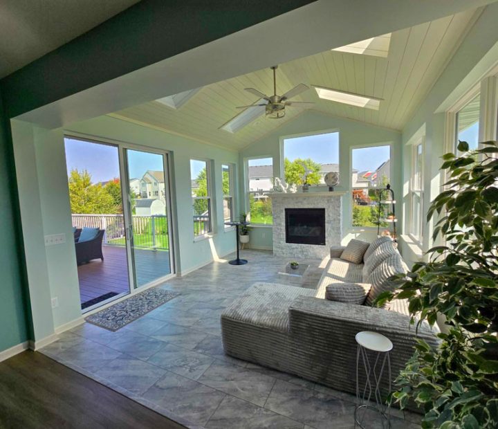A bright sunroom with light blue walls, a stone fireplace, vaulted ceiling with skylights, and large sliding glass doors opening to a wooden deck.